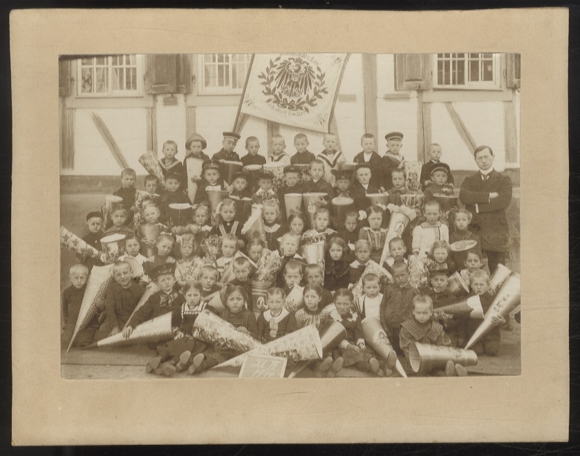 Fotograficzne zdjęcie grupowe dzieci z rożkiem cukrowym przed budynkiem szkoły, nauczyciel, flaga, 1914