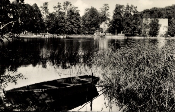 Postcard Lindow in der Mark, calm lake, boat in the foreground, trees and buildings in the background