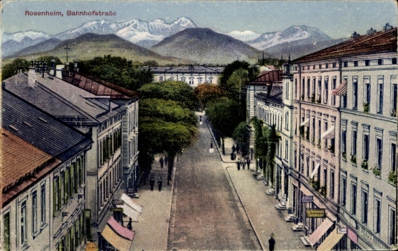 Postcard Rosenheim Oberbayern, Bahnhofstraße. A street with trees, houses and mountains in the background...