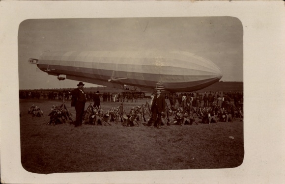 Photo Postcard Zeppelin airship over an airfield, onlookers, mounted rifles and spiked helmets