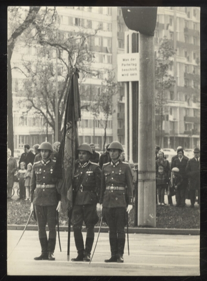 Photograph of GDR soldiers in uniform, flag, slogan board for the party congress