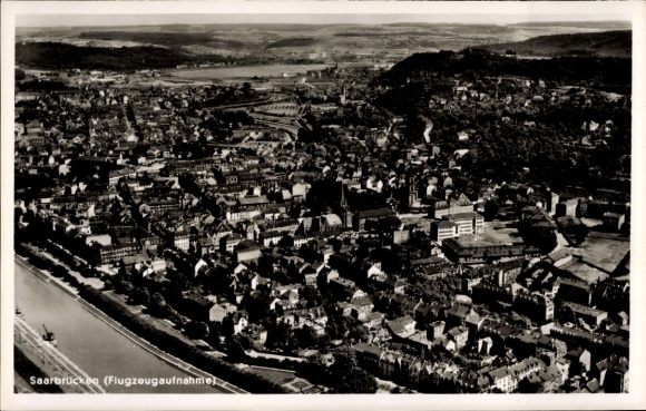 Ak Saarbrücken im Saarland, Luftaufnahme von  Fluss im Vordergrund, Stadtansicht, Berge im Hinter